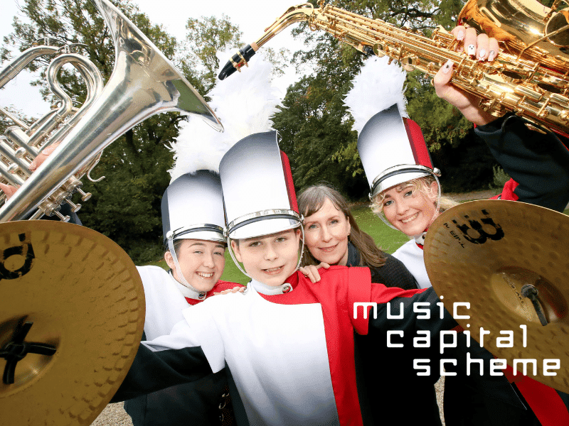 Three Band Members In Uniform Pose With Their Instruments A Young Woman With A Tuba A Young Boy With Symbols And Another Young Women With A Saxphone They Are Also Joined By Another Woman. 2.png